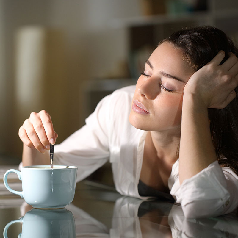 woman falling asleep at table while stirring liquid in mug